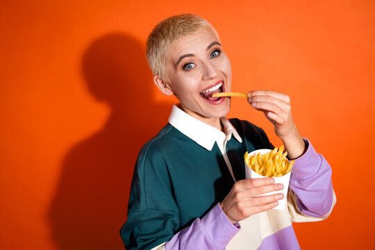 Young cheerful woman enjoying a snack of crispy French fries on a vibrant orange background, embracing casual style