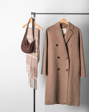 Brown wool coat or jacket, beige alpaca scarf and dark suede handbag on hanger against wall background. Warm wool clothing.