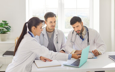 Fototapeta premium Young group of confident doctors sitting with laptop at the desk in clinic during a meeting and discussing diagnosis, checking test results or planning treatment. Medicine ad healthcare concept.