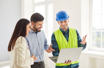Smiling construction worker in blue hard hat and safety vest showing information on laptop to young...
