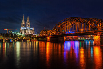 Fototapeta premium As dusk descends over Cologne, the blue hour transforms the city into an enchanting spectacle. In this photograph, the Hohenzollernbrücke, with its robust arches and love locks, forms a powerful foreg