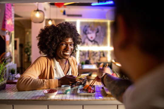 Young african woman taking a photo with her smart phone of the sushi a chef prepared in a sushi bar