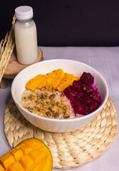A healthy oatmeal bowl topped with fresh mango slices, diced dragon fruit, and crunchy granola, served with a bottle of milk. A nutritious breakfast with vibrant colors and natural ingredients.