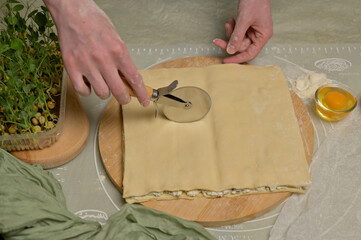 Square sheet of dough cut into strips with woman's hands on wooden board. Cutter, egg, green towel, microgreens sprouts on kitchen table. Home cooking baking puffs