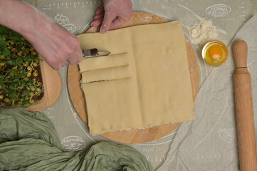 Top view square sheet of dough cut into strips with woman's hands on wooden board. Cutter, egg, green towel, microgreens sprouts on kitchen table. Home cooking baking puffs