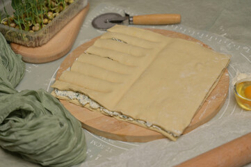 Square sheet of dough cut into strips on wooden board. Cutter, egg, green towel, microgreens sprouts on kitchen table. Home cooking baking puffs