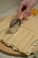 Close up square sheet of dough cut into strips with woman's hands on wooden board. Cutter, egg on kitchen table. Home cooking baking puffs