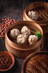 Siomay a traditional steamed fish dumpling dish served in a bamboo steamer with lime and peanut sauce. The setting features raw peanuts and spices on a dark rustic background.