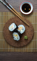 Three sushi rolls with crab, cucumber, and cream cheese on a cork mat, served with wasabi, soy sauce, and chopsticks on a bamboo mat. A perfect Japanese meal presentation.