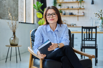 Mature confident businesswoman with notepad sitting in chair, looking at camera