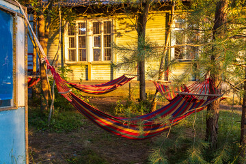 Two colorful striped hammocks hanging between trees near a yellow wooden house at sunset.