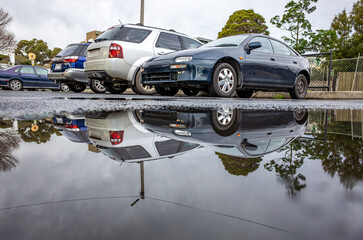 A row of parked cars is reflected in a large puddle on wet asphalt after rainfall or storm, indicating water accumulation and drainage issues in an urban city or parking lot. © Doublelee
