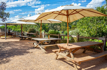 A sunny outdoor seating area with wooden picnic tables and large beige umbrellas in a garden. A shaded outdoor dining or resting space typical of parks, wineries, or casual restaurant patios.