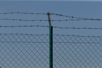 The image depicts a barbed wire fence set against a bright blue sky, creating a stark contrast that...