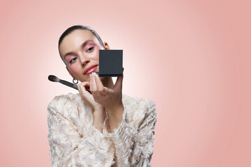A young woman in a cream textured jacket applying blush with a makeup brush while holding a compact...
