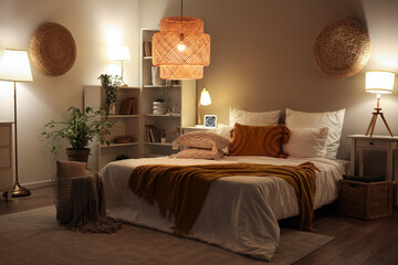 Interior of cozy bedroom with comfortable bed, blanket and glowing lamps at night