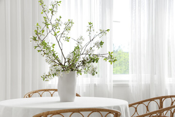 Vase with blooming branches on table near window in dining room