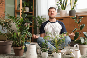 Young handsome man meditating among plants in room