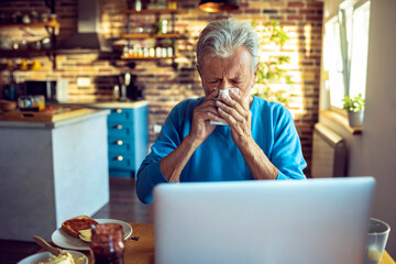 Senior man having a headache at home and blowing his nose while having breakfast
