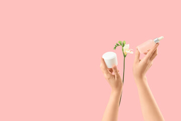 Female hands with bottles of cosmetic products and beautiful flowers on pink background