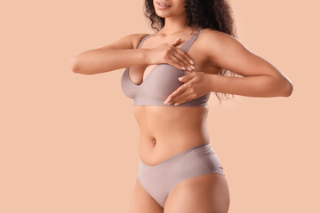 Young African-American woman in underwear checking her breast on beige background. Cancer awareness...