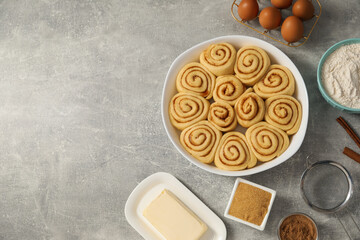 Raw cinnamon rolls in baking dish and different ingredients on grey table, flat lay. Space for text
