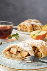 Pieces of tasty apple strudel with powdered sugar and fork on white table against grey background, closeup