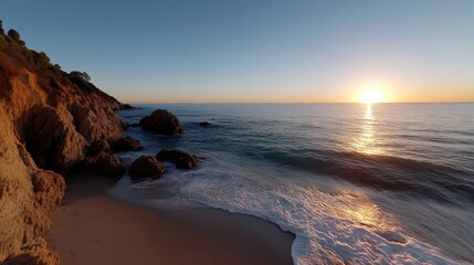Obraz premium Coastal sunset scene with rocks, waves, and sandy beach. Golden light bathes the shore as the sun dips below the horizon. Calm ocean meets the rugged cliff edge