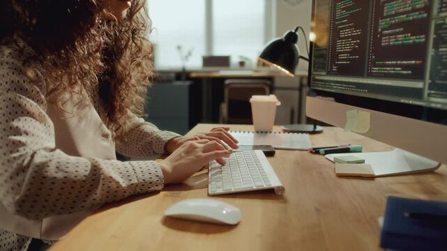 Young female developer typing on wireless keyboard and examining data on monitor while working on coding project at computer desk in modern office