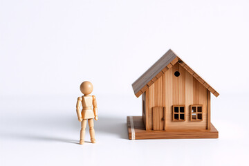 A small wooden mannequin stands beside a simple miniature wooden house against a plain white backdrop, creating a minimalist and conceptual scene.