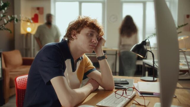 Time lapse of apathetic young man resting head on hand and staring boredly at computer monitor while sitting at desk in modern office, with coworkers moving in background