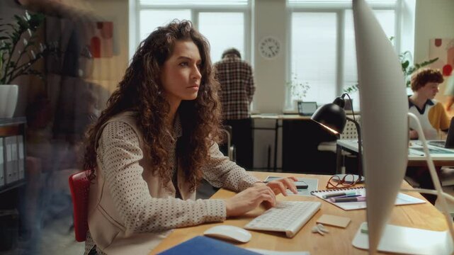 Time lapse of young beautiful businesswoman in casual outfit sitting at desk,typing on wireless keyboard as working on computer during day in office, colleagues walking in background