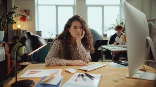Time lapse of young bored businesswoman with curly hair resting head on hand, sitting at desk in modern open plan office, looking apathetically at computer screen, with coworkers walking in background