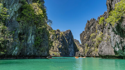 Tourist canoes sail along the turquoise lagoon surrounded by sheer cliffs. Green tropical vegetation on steep rocky slopes. The blue sky. Philippines. Palawan. Big Lagoon. The Bacuit archipelago.