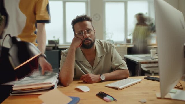 Overworked middle-eastern man with glasses resting his head on hand, looking tiredly into camera while sitting at desk in modern office with pile of papers nearby. Time lapse, video portrait