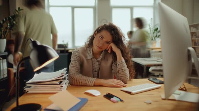 Portrait of young frustrated woman resting head on hand and looking into camera while sitting at desk in open plan office, overloaded with paperwork stacked nearby. Time lapse