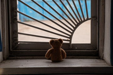 Children's brown bear sitting on a window with bars and looking out the window in an old abandoned...