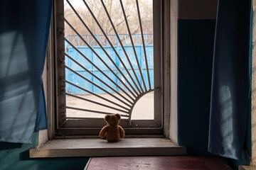 Children's brown bear sitting on a window with bars and looking out the window in an old abandoned...