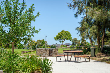 A picnic table and a stone barbecue in a park, surrounded by lush trees and green outdoor space. Communal amenities for social gatherings in a public park in the suburb of Tarneit, Australia