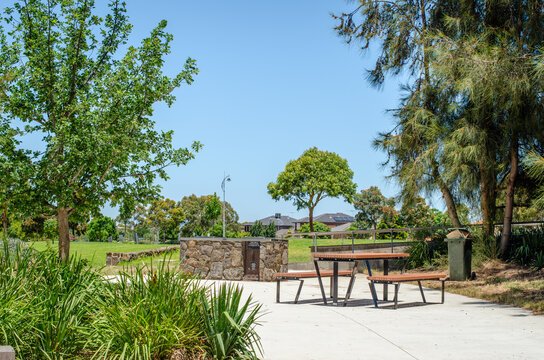 A picnic table and a stone barbecue in a park, surrounded by lush trees and green outdoor space. Communal amenities for social gatherings in a public park in the suburb of Tarneit, Australia