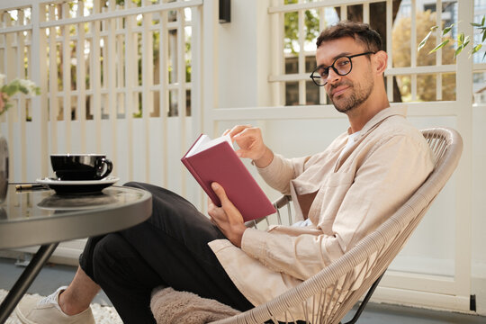 A young man sitting comfortably in a chair on a patio, reading a book and enjoying a cup of coffee. The outdoor setting creates a relaxed atmosphere.