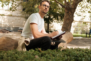 Obraz premium A thoughtful man with glasses sits on a log in a park, diligently writing in his notebook, surrounded by greenery and sunlight. The scene conveys creativity, reflection, and relaxation.