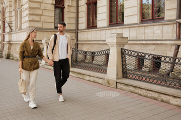 A couple strolls happily hand in hand down a charming historic street, enjoying a leisurely day out together. The cityscape adds to the picturesque scene of their romantic walk.