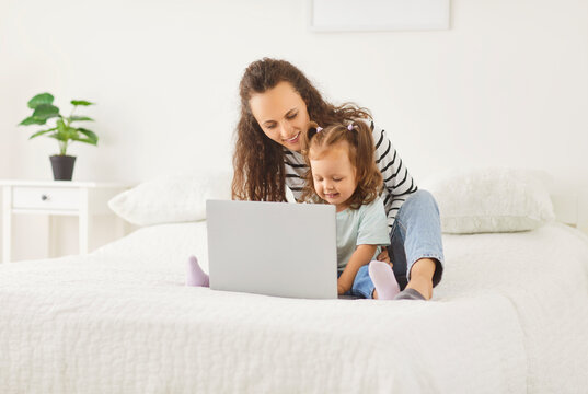 Mother and young daughter resting, relaxing together on a bed at home, sharing online entertainment in internet using laptop. Cozy leisure moment that highlights family bonding and modern pastimes.