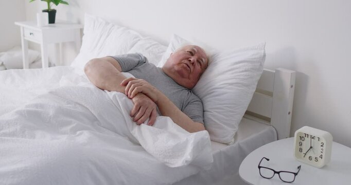 Upset sleepless senior elderly man lying on bed in bedroom under white blanket at home in bad thoughts. Retired person cant sleep in the morning indoors. Insomnia concept. 4k video.