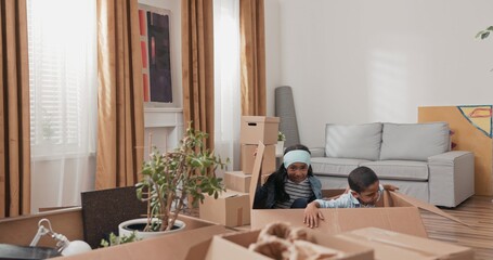 A sister and her younger brother play inside empty cardboard boxes while moving. Surrounded by packed items, they laugh and hide, enjoying joyful childhood moments in their new apartment.