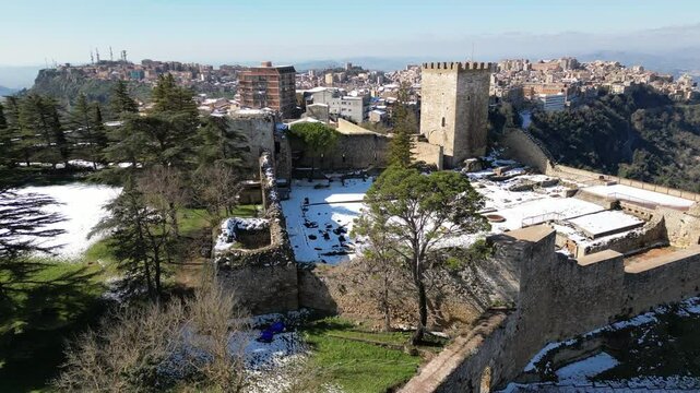 Flying over trees in courtyard of massive castle towards small village of Enna, located in mountains of central Sicily Italy
