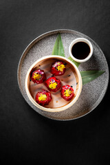 A top view of a bamboo steamer filled with vibrant red dumplings garnished with yellow petals, accompanied by a small bowl of soy sauce on a textured black background.