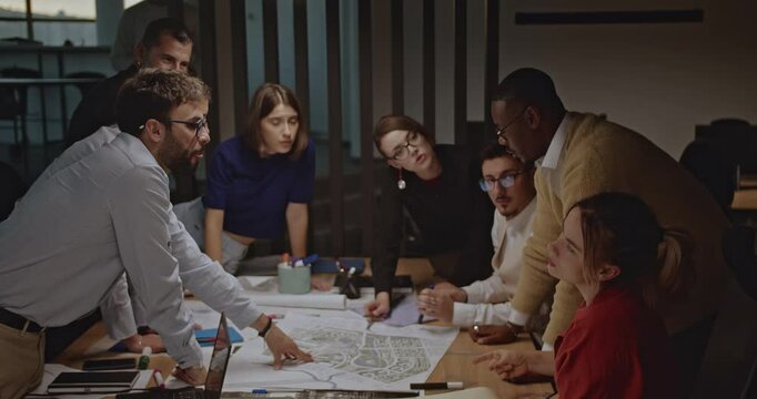 Diverse team of business people in a night meeting discussing strategy around a table with blueprints.
