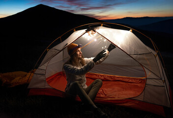 Shot of a young woman wearing gloves and an orange hat, adding lights to a tent in the mountains at...